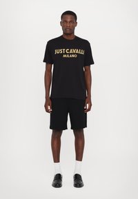Černé bavlněné tričko s zlatým potiskem "JUST CAVALLI MILANO", v kombinaci s černými šortkami, bílými ponožkami a černými mokasíny. Jednoduchý, moderní design.