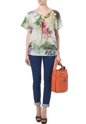 Blusa a maniche corte con motivo floreale nei toni del verde, rosa e viola; abbinata a jeans skinny blu scuro e una borsa arancione.