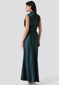 Donna in un abito satinato senza maniche di un verde scuro, con colletto alto e una cintura, che indossa orecchini e tacchi alti beige, rivolta di lato su uno sfondo bianco.