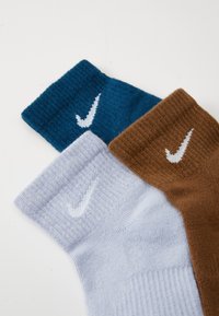 Drei Paar Socken in Blau, Hellgrau und Braun, mit gerippten Bündchen und weißen Nike-Swoosh-Logos auf jeder Socke.