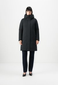 Zwarte gewatteerde jas met een hoge kraag en verstelbare capuchon, voorzien van een ritssluiting en zijzakken; gedragen over donkere jeans en zwarte hakken.