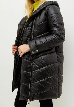 Mujer que lleva una chaqueta larga acolchada negra con capucha, desabrochándola para mostrar un suéter de cuello alto de color amarillo neón debajo.