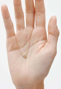 Collar de oro con dos encantos en forma de corazón entrelazados sostenidos en una palma abierta, con una cadena de eslabones fina y superficie lisa y pulida.