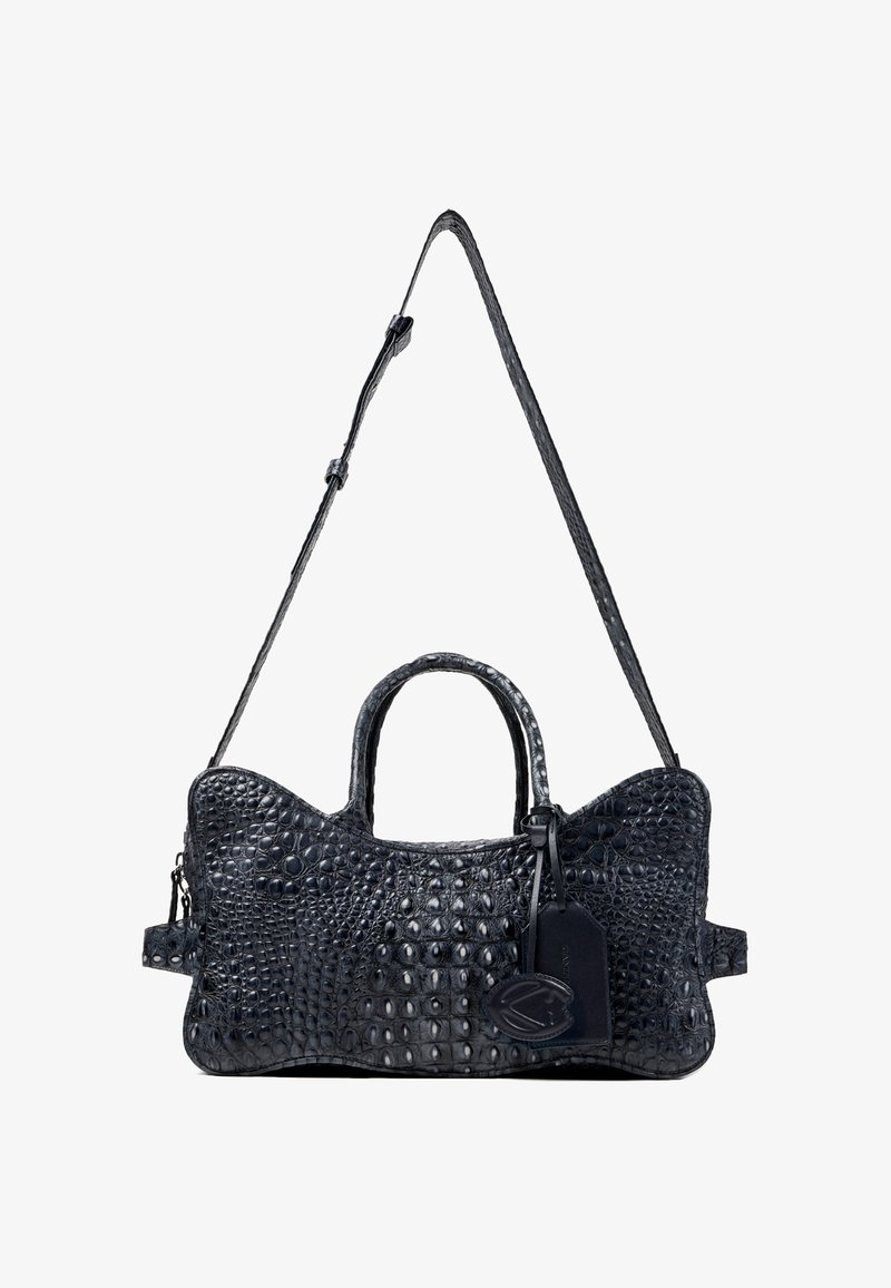 Sac à main en cuir noir texturé avec bandoulière ajustable, deux poignées et étiquette à bagages attachée avec emblème décoratif.