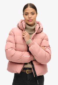 Giacca a vento rosa con cappuccio, caratterizzata da un design trapuntato, polsini elasticizzati e un dettaglio logo ben visibile. Indossata sopra un dolcevita beige lavorato a maglia.