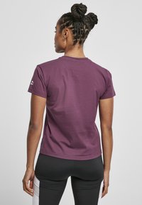 Femeie cu părul împletit în două cocuri, îmbrăcată cu un tricou scurt cu mânecă de culoare purpurie și colanți negri cu panouri laterale albe, stând în fața unui fundal simplu.