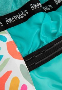 Turkusowa tkanina z czarną taśmą z logo, z kolorowymi abstrakcyjnymi wzorami. Zawiera gładki zamek błyskawiczny i miękki materiał o teksturowanej powierzchni.