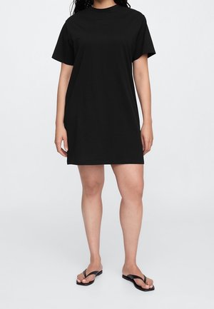 Frau trägt ein kurzärmliges schwarzes T-Shirt-Kleid und schwarze Flip-Flops, steht vor einem einfarbigen hellgrauen Hintergrund.