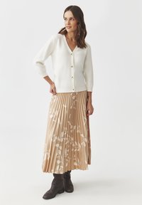 White, knitted cardigan with a V-neck and three-quarter sleeves. Paired with a pleated, beige skirt featuring a white leaf pattern and brown ankle boots.