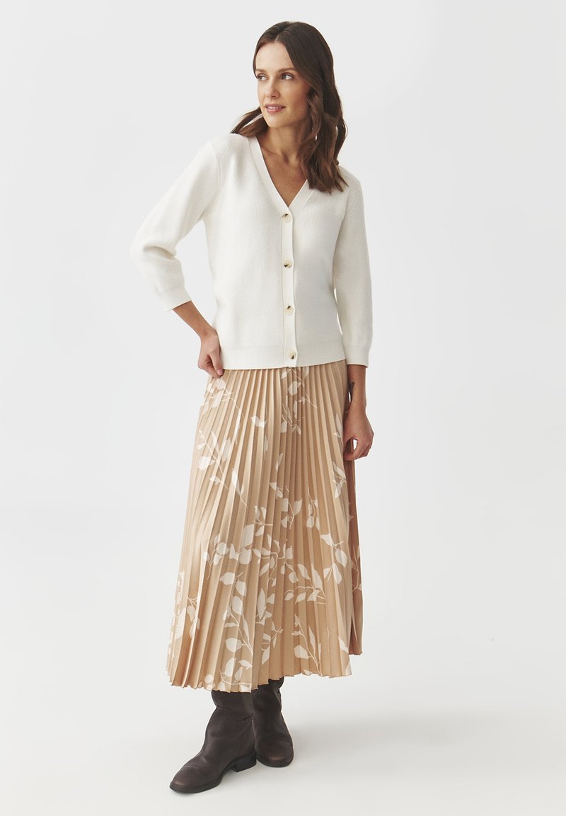 Biały, dziergany kardigan z dekoltem w serek i rękawami do trzech czwarte. Połączony z plisowaną, beżową spódnicą z białym wzorem liści oraz brązowymi botkami za kostkę.