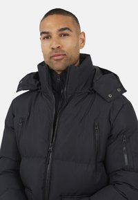 Zwarte gewatteerde jas met een hoge kraag, ritszakken en een gestructureerd oppervlak. Beschikt over een afneembare capuchon en duurzame ritsen.