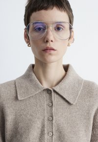 Beige Wollcardigan mit kragenförmigem Ausschnitt, fünf Knöpfen auf der Vorderseite und strukturiertem Strick. Getragen mit runden, klaren Brillen und goldenen Creolen.