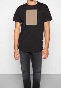 Czarna koszulka z krótkim rękawem z pomarańczowym geometrycznym logo na beżowym tle, z okrągłym dekoltem i luźnym krojem. Połączona z wyblakłymi czarnymi dżinsami.