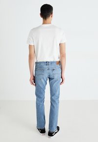 Világoskék denim farmer, egyenes lábadról készült, hátsó zsebekkel és barna bőr címkével a derékrészen. Fehér pólóval viselve.