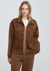 Giacca marrone con zip, dal texture scamosciato, polsini a costine e un piccolo logo sul petto. Abbinata a pantaloni marroni coordinati.