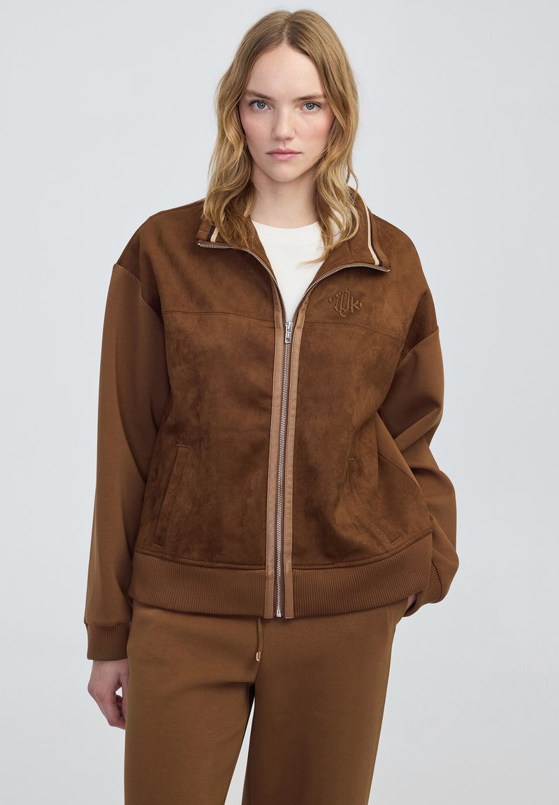 Giacca marrone con zip, dal texture scamosciato, polsini a costine e un piccolo logo sul petto. Abbinata a pantaloni marroni coordinati.