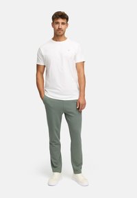 Ung mand står med hænderne i lommerne, iført hvid t-shirt, olivenfarvede bukser og hvide sneakers foran en ensfarvet baggrund.