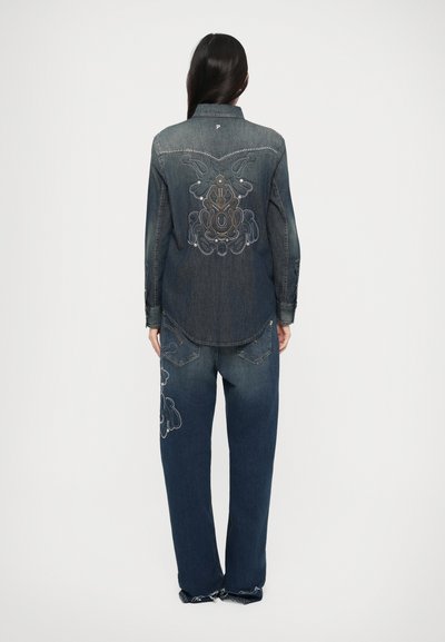 Denim overhemd en broek set in donkerblauw. Het overhemd heeft geborduurde details op de rug; de broek heeft vergelijkbare borduursels op de benen.