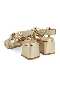 Sandalias trenzadas de oro con un tacón cuadrado y grueso. Presentan un acabado metálico suave con un diseño de tira texturizada y acentos dorados.