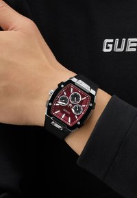 Poignet avec montre à bracelet noir, cadran hexagonal rouge avec trois sous-cadrans et index argentés sur une manche noire.