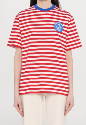 Personne portant un t-shirt à manches courtes rayé rouge et blanc avec un col bleu et un logo circulaire bleu sur le côté gauche de la poitrine.