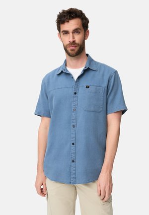 Homme portant une chemise à manches courtes bleu clair boutonnée par-dessus un t-shirt blanc et un pantalon cargo beige, debout contre un fond clair uni.