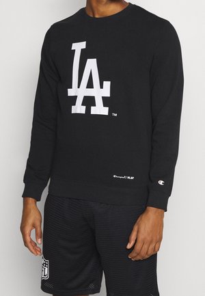 Man med svart långärmad sweatshirt med stort vitt "LA"-logotyp och svarta mesh-shorts med en vit NFL-logotyp på vänster ben.