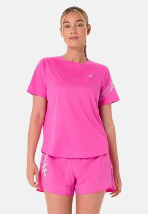 Mujer vistiendo camiseta deportiva rosa brillante y shorts a juego con detalles de logotipo blanco, de pie contra un fondo claro y liso.