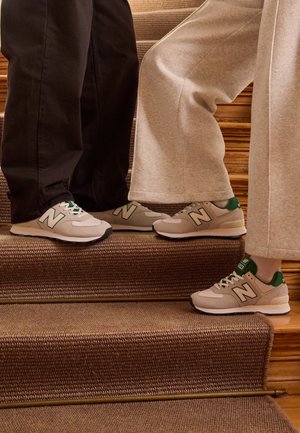Deux personnes portant des baskets New Balance beige et vert se tiennent sur des escaliers moquettés, un pied levé sur une marche plus haute.