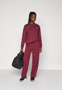 Maroon sweatshirt with drawstring and logo, paired with matching maroon pants. Black sports bag in hand. Smooth fabric and casual fit.