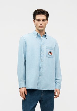 Giovane uomo con camicia di jeans azzurro chiaro con bottoni e logo Tommy Hilfiger sulla tasca sul petto e jeans blu scuro.