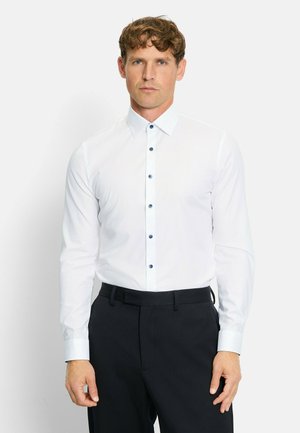 Weißes Langarmhemd mit klassischem Kragen, mit dunkelblauen Knöpfen entlang der Knopfleiste, kombiniert mit dunkler Hose.