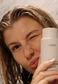 Bouteille blanche étiquetée "OUAI" tenue dans une main, avec une finition lisse. L'arrière-plan présente une texture marbrée ; les cheveux sont mouillés et ébouriffés.