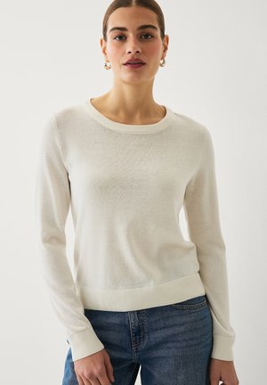 Woman wearing a cream long-sleeve crewneck sweater and blue jeans, standing against a plain light background.