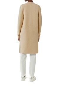 Osoba ubrana w długi beżowy sweter, białe spodnie i białe trampki z srebrnymi obcasami, stojąca tyłem na białym tle.