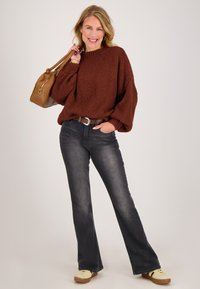 Bruine gebreide trui met structuur, donker uitlopende spijkerbroek, beige sneakers en een bruine handtas met een ronde metalen sierlijke accenten. Model staat lachend.