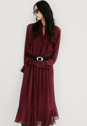 Mujer de cabello largo y oscuro, con gafas de sol oscuras, vistiendo un vestido burdeos fluido con volantes y un cinturón negro, de pie contra una pared blanca.