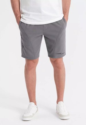 Pantaloni corti grigi in tessuto sintetico, con una texture liscia, tasche laterali e un logo discreto sull'orlo. Abbinati a una maglietta a collo rotondo.