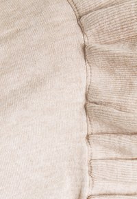 Smėlio spalvos megztinis iš megztos medžiagos su ribotu raštu ir surinktu detalumu vienoje pusėje, demonstruojantis minkštą audinį ir subtilų spalvų niuansą.