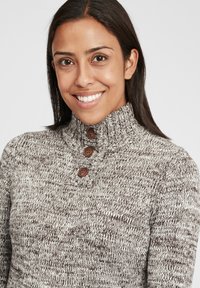 Strikkegenser i nyanser av beige og brunt, med høy krage og tre brune knapper ved halsen. Strukturer mønster gjennom hele.