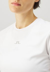 Bílý sportovní tričko s kulatým výstřihem a stříbrným logem uprostřed. Hladká textura, přiléhavý design, minimalistická estetika.