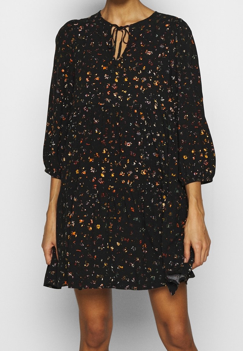 Femme portant une mini robe noire à manches longues, à motif floral, avec un col à nouer et une jupe froncée.