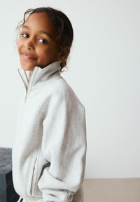 Sweat-shirt zippé gris clair en tissu doux, avec un col montant et une coupe décontractée. Couture visible et détail de poche latérale.