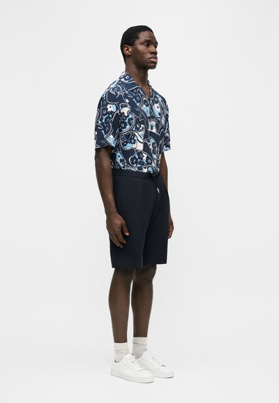 Homme debout portant une chemise bleue à manches courtes avec un motif floral, un short bleu marine, des chaussettes blanches et des baskets blanches sur un fond uni.