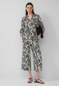 Zwart-wit blouse met bladraster en v-hals, gecombineerd met wijde, verkorte broek. Textuurstof met zakken, gedragen met platte zwarte sandalen.