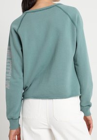 Turkusowy sweter z rękawami raglanowymi, ściągaczami i widocznymi szwami. Napis "AND ETCH NY" w poziomie na lewym rękawie. Casualowy krój i design.