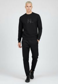 Sudadera negra con un logo sutil en el frente, combinada con pantalones negros a juego y zapatillas negras. Tejido liso, diseño ajustado.
