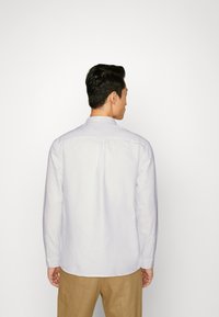 Camisa de manga larga lila claro con cuello abotonado, que presenta un pliegue en la parte trasera y una textura sutil. Combinada con pantalones color tostado.