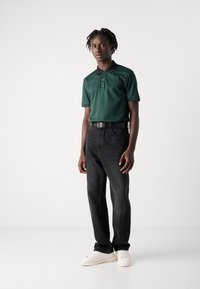 Mladý muž s tmavou pokožkou a krátkymi dredmi je oblečený v zelenej polokošeli, čiernych džínsoch, bielych teniskách a čiernom opasku, stojí na bielom pozadí.