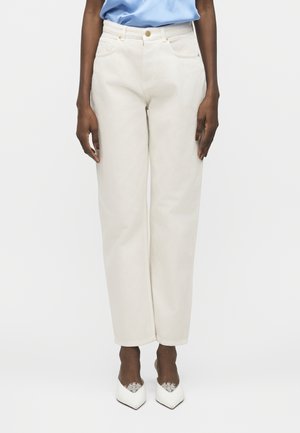 Personne portant un jean blanc taille haute à jambes droites, une chemise bleu clair rentrée, et des chaussures blanches pointues avec un bord en dentelle.